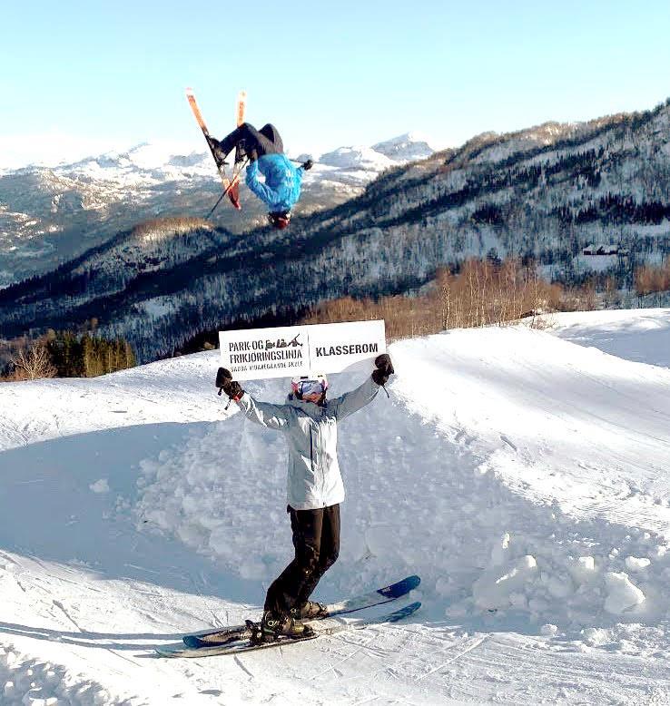 Ein person gjer salto på ski over eit hopp, medan ein peron i forgrunnen held opp eit skilt der det står «Park og frikjøringslinja Sauda vidaregåande skule | Klasserom». - Klikk for stort bilde